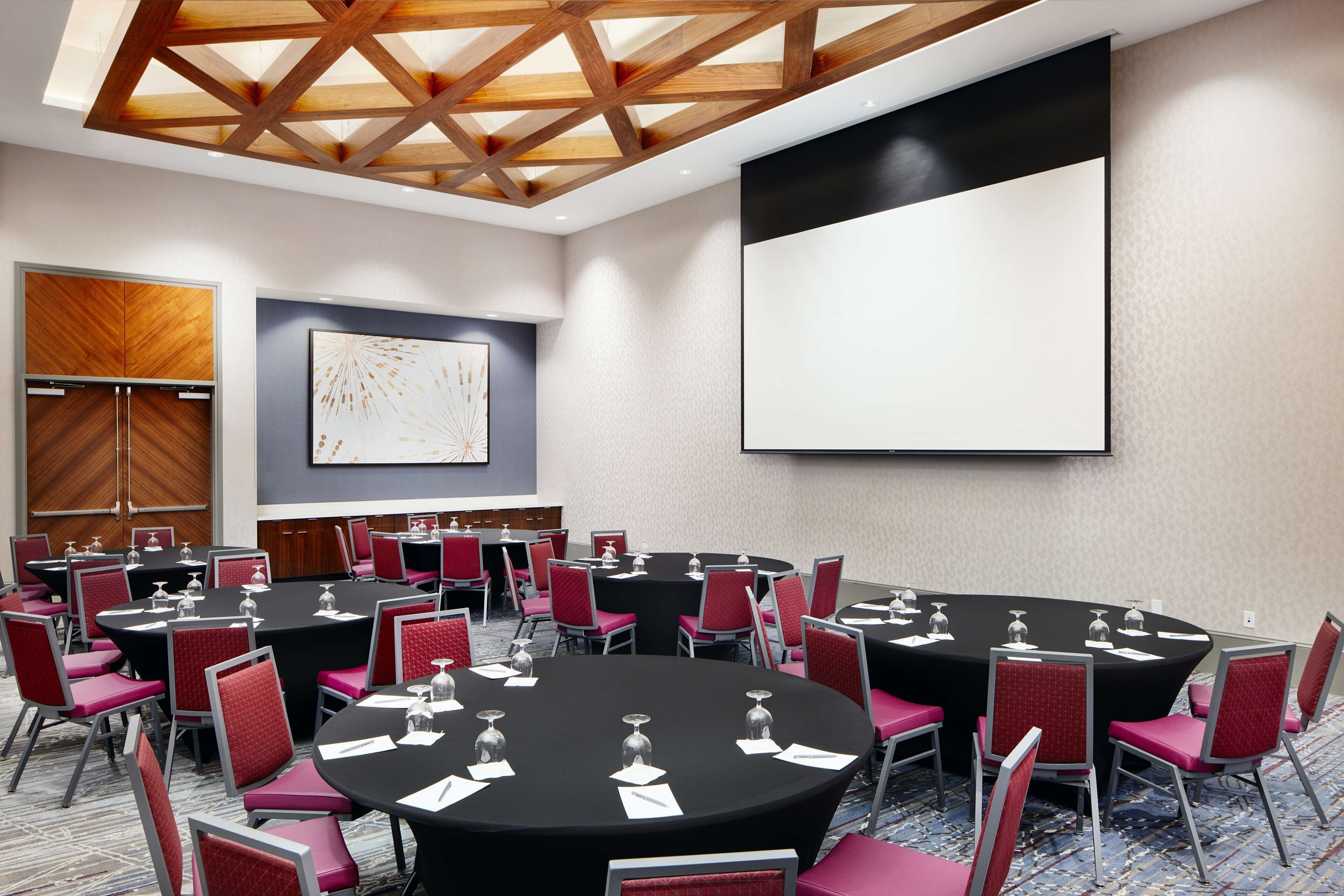 A conference room with multiple tables and chairs, a large screen, and a wooden ceiling.