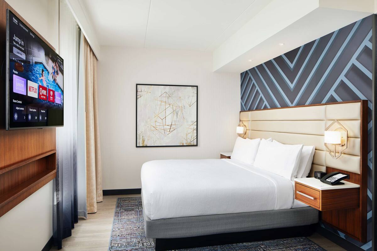 Een hotelkamer met een grote flatscreen televisie aan de muur, een bed met hoofdeinde, nachtkastjes en een lijst met een abstract schilderij.