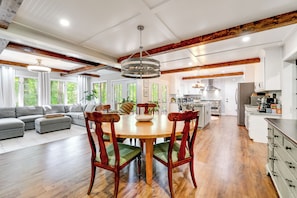 A spacious kitchen with wooden beams, a dining area with a round table and chairs, and a living room with a sofa and large windows.