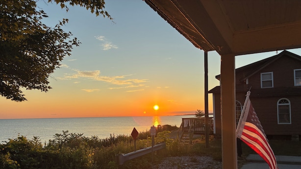 A sunset view from a porch overlooking a body of water, with a wooden house and an American flag in the foreground.