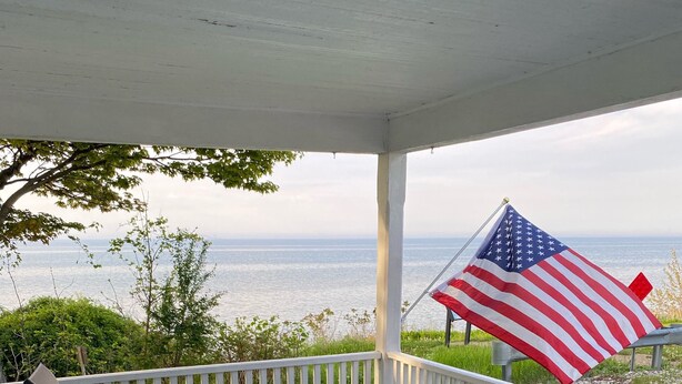 Um alpendre coberto com mesa e cadeiras, uma bandeira americana e vista para o oceano.