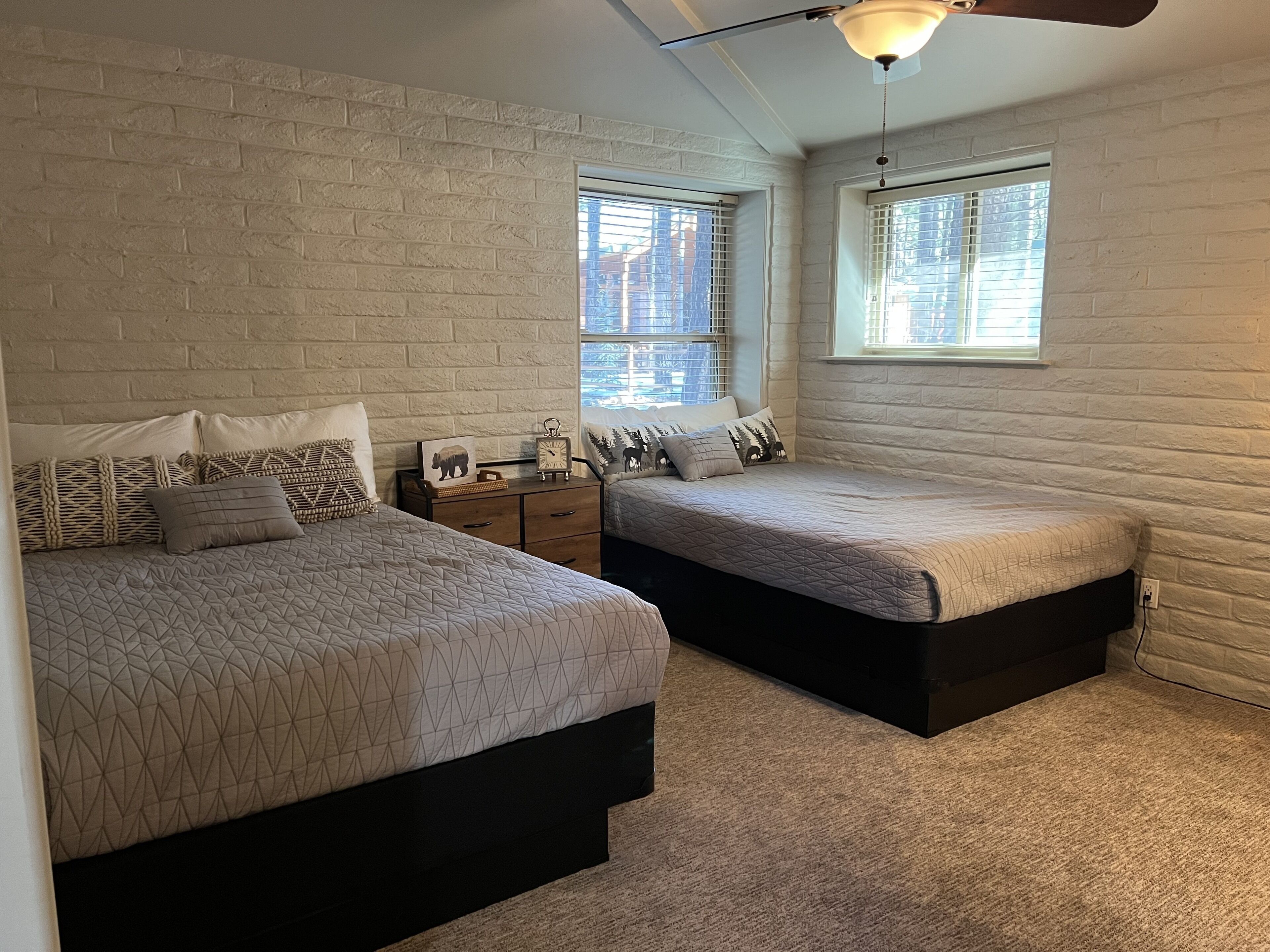 A bedroom with two beds, a white brick wall, a ceiling fan, and windows with blinds.