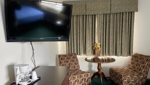 A hotel room with a flat-screen TV, a coffee maker, two patterned chairs, a round table with a vase, and a large window with curtains. - Heritage inn (Watertown)