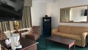 A living room with a flat-screen TV, a coffee machine, a sofa, a patterned armchair, and a small refrigerator. - Heritage inn (Watertown)