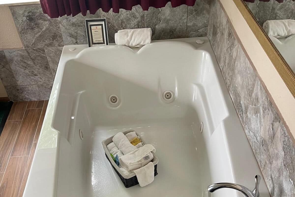 A large, rectangular bathtub with a wooden base, a built-in showerhead, and a small shelf with towels.