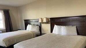 A hotel room with two beds, each with a wooden headboard and white bedding. There is a nightstand with a lamp between the beds. - Heritage inn (Watertown)
