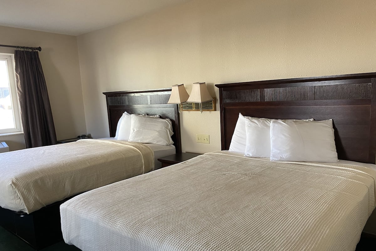 A hotel room with two beds, each with a wooden headboard and white bedding. There is a nightstand with a lamp between the beds.