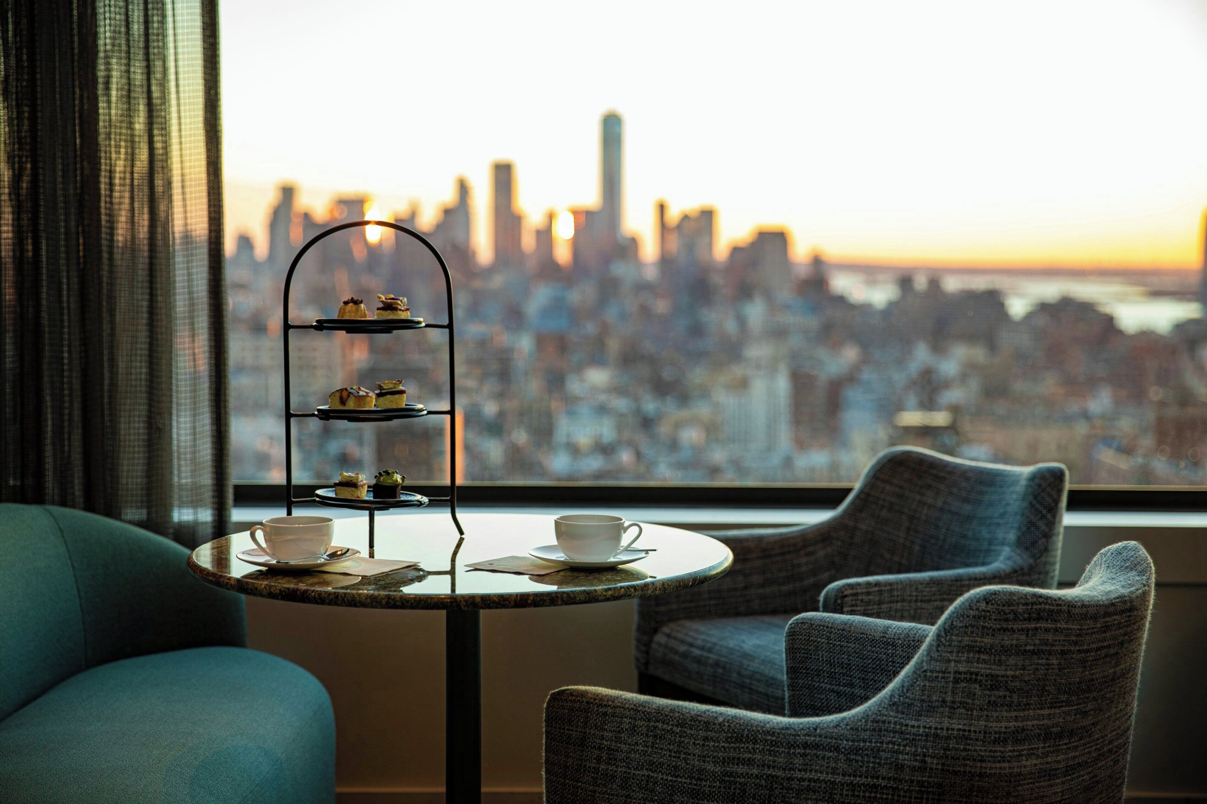 Une pièce avec vue sur la ville, une petite table ronde surmontée d’un présentoir à gâteaux, deux chaises et un canapé.