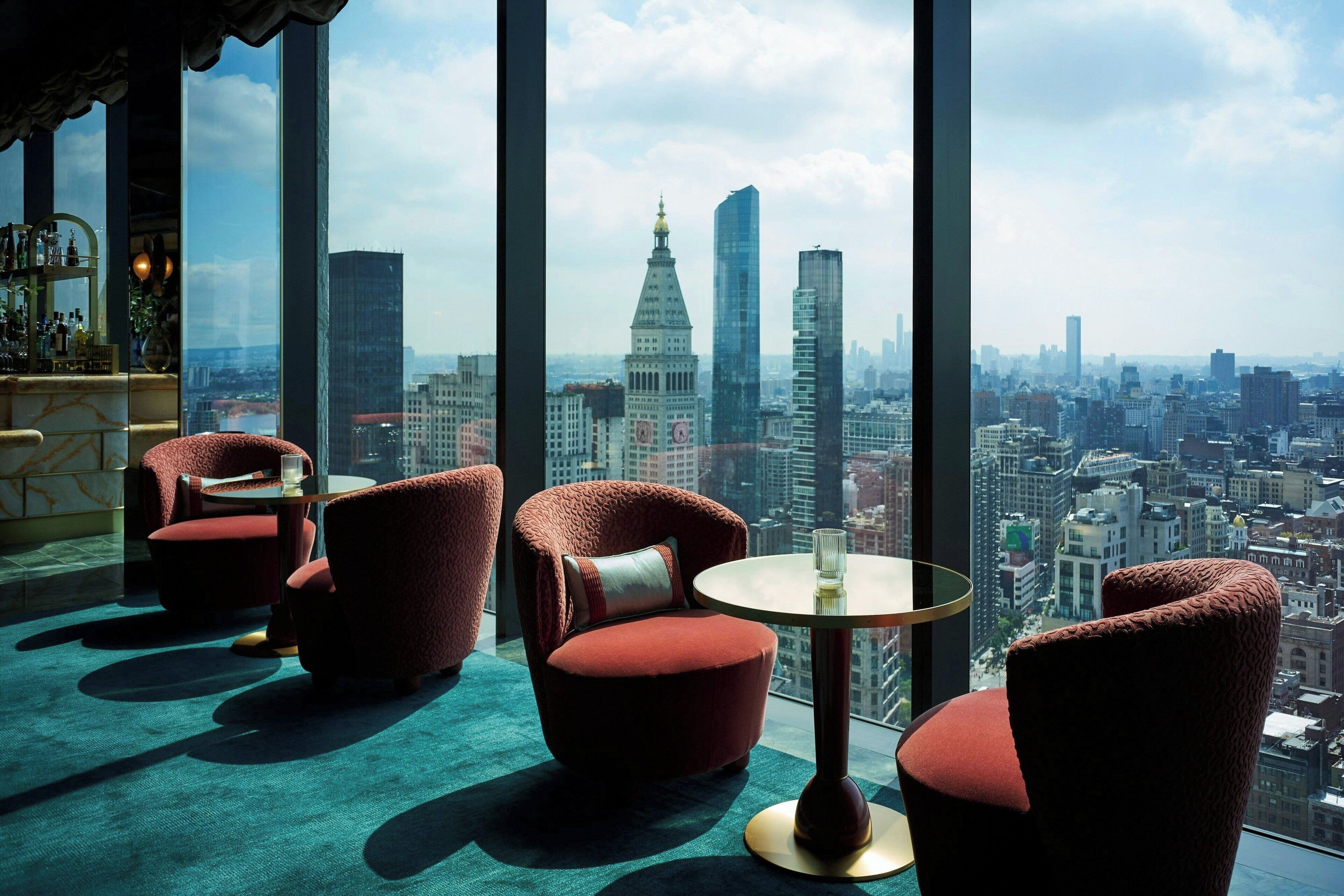 Habitación en un rascacielos con vistas panorámicas de la ciudad, que incluye sillas tapizadas en rojo, una mesa redonda y una zona de bar con botellas.