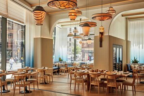 A spacious cafe with wooden tables and chairs, large windows offering a view of the street, and multiple hanging pendant lights.