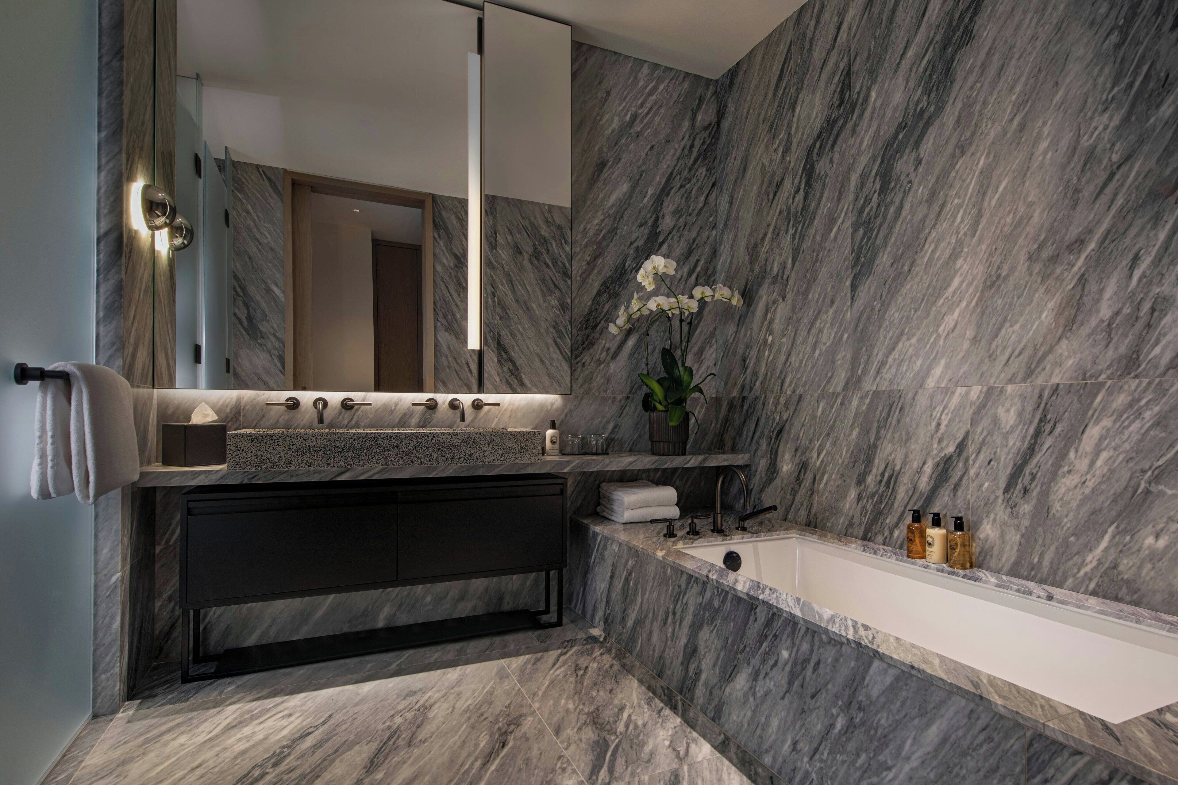 Une salle de bain moderne avec un mur en marbre, un grand miroir, un meuble-lavabo, une baignoire et un pot d’orchidée.