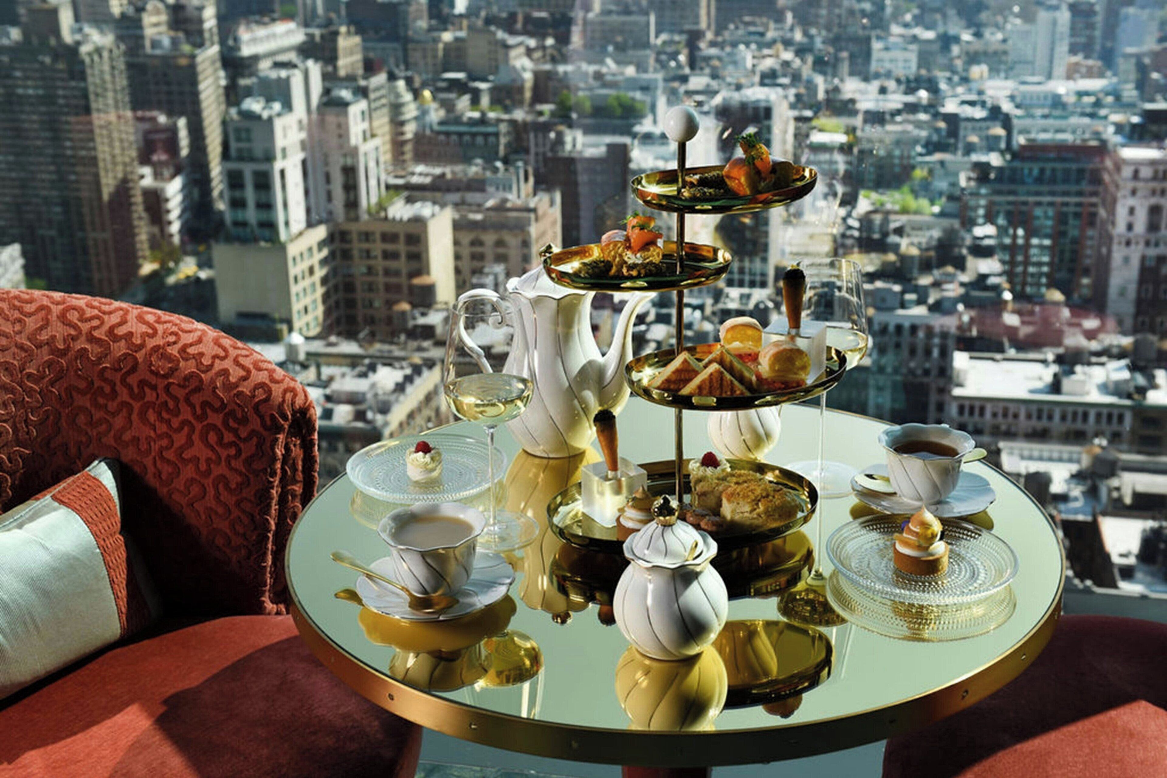 Una mesa con un tres niveles de bandeja de comida y bebidas, una tetera y una taza, frente a una vista panorámica de la ciudad.