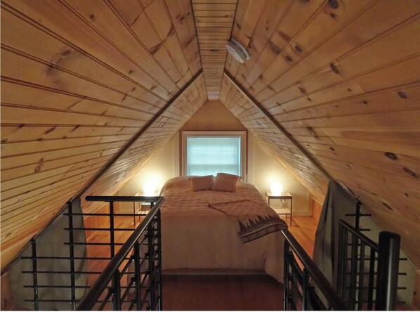 Cozy attic retreat with a sloped ceiling, wooden beams, and a small window, featuring a comfortable bed and a nightstand with a lamp.