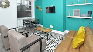 A modern living room with teal walls, a black and white patterned rug, a brown sofa with a yellow pillow, a wooden coffee table, and a TV mounted on the wall. - Townhouse @ 202 Walthall Street Crewe (Crewe)
