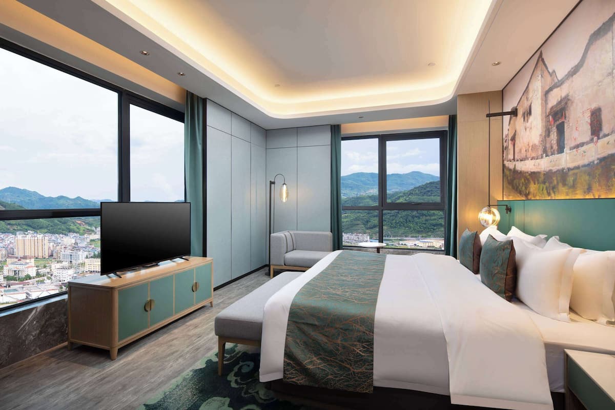 Habitación de hotel moderna con una cama grande, televisión, zona de estar y vistas a la ciudad a través de amplios ventanales.