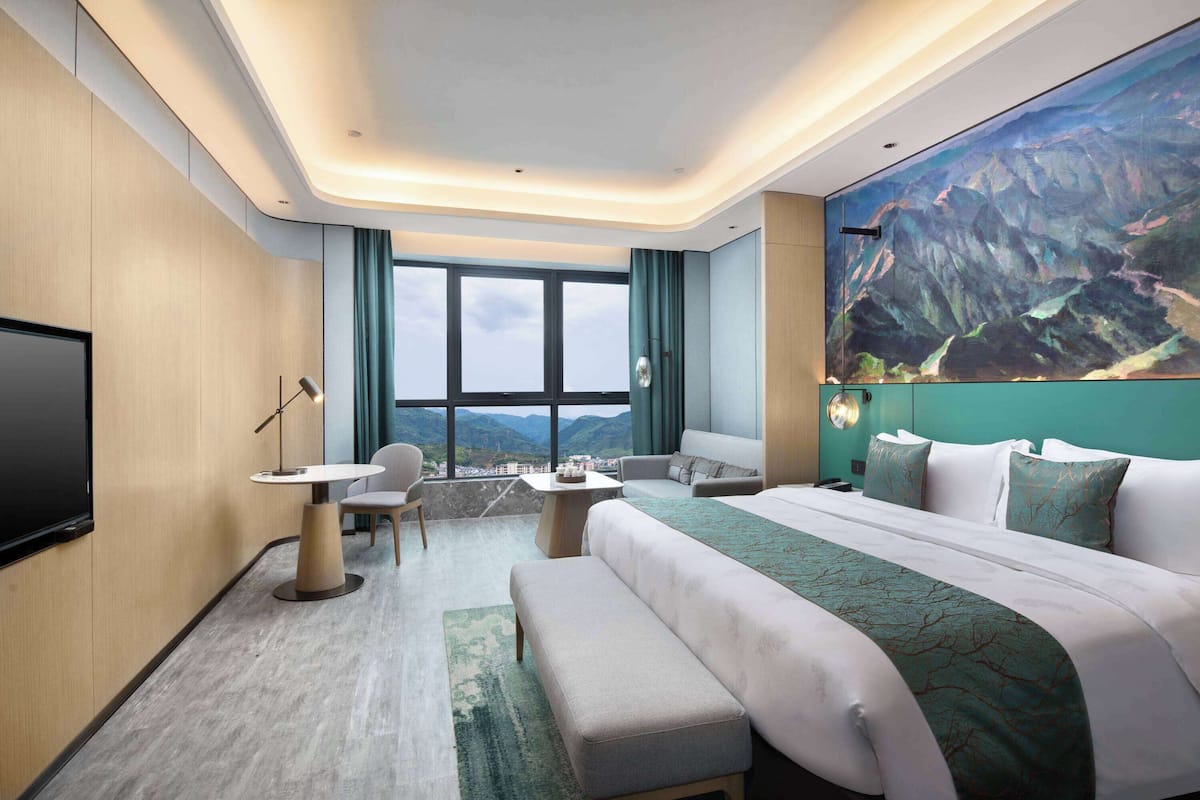 Habitación de hotel moderna con una cama grande, zona de estar con mesa y sillas, televisión de pantalla plana y un ventanal con vistas a la montaña.