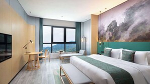 Una habitación de hotel moderna con una cama grande, un escritorio y una silla. Hay un cuadro en la pared y vistas a las montañas a través de la ventana.