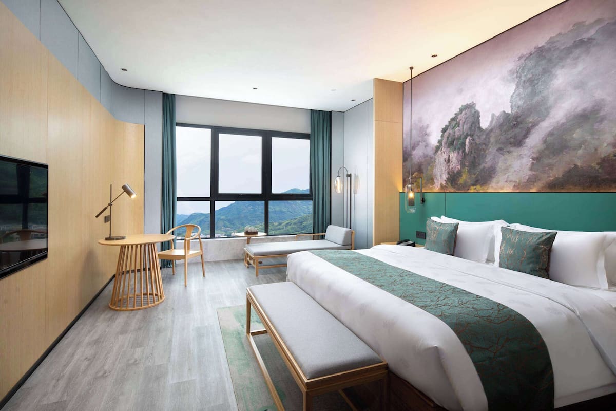Una habitación de hotel moderna con una cama grande, un escritorio y una silla. Hay un cuadro en la pared y vistas a las montañas a través de la ventana.