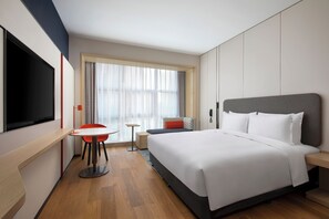 A modern hotel room with a large bed, a flat-screen TV, a small table, and a window with curtains.