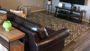 Ein Wohnzimmer mit einem dunkelbraunen Ledersofa, einem Holztisch, einem Flachbildfernseher und einem gemusterten Teppich.