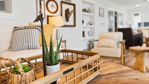 Una sala de estar acogedora con una mesa de centro de madera, un sofá blanco y un cojín a rayas. Se observan estanterías con adornos y una lámpara.