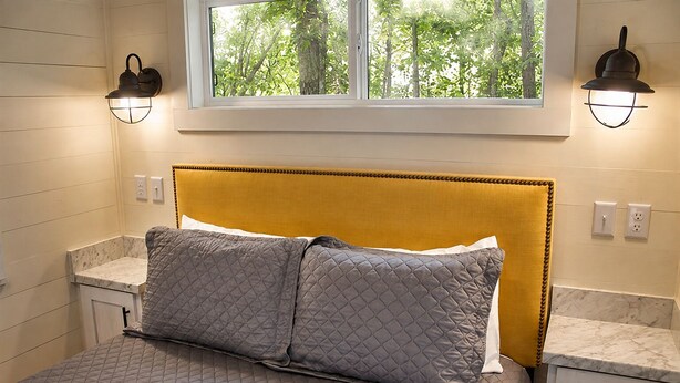 Un dormitorio con cama, dos mesitas de noche, una ventana con vista a árboles y luces fijadas a la pared.
