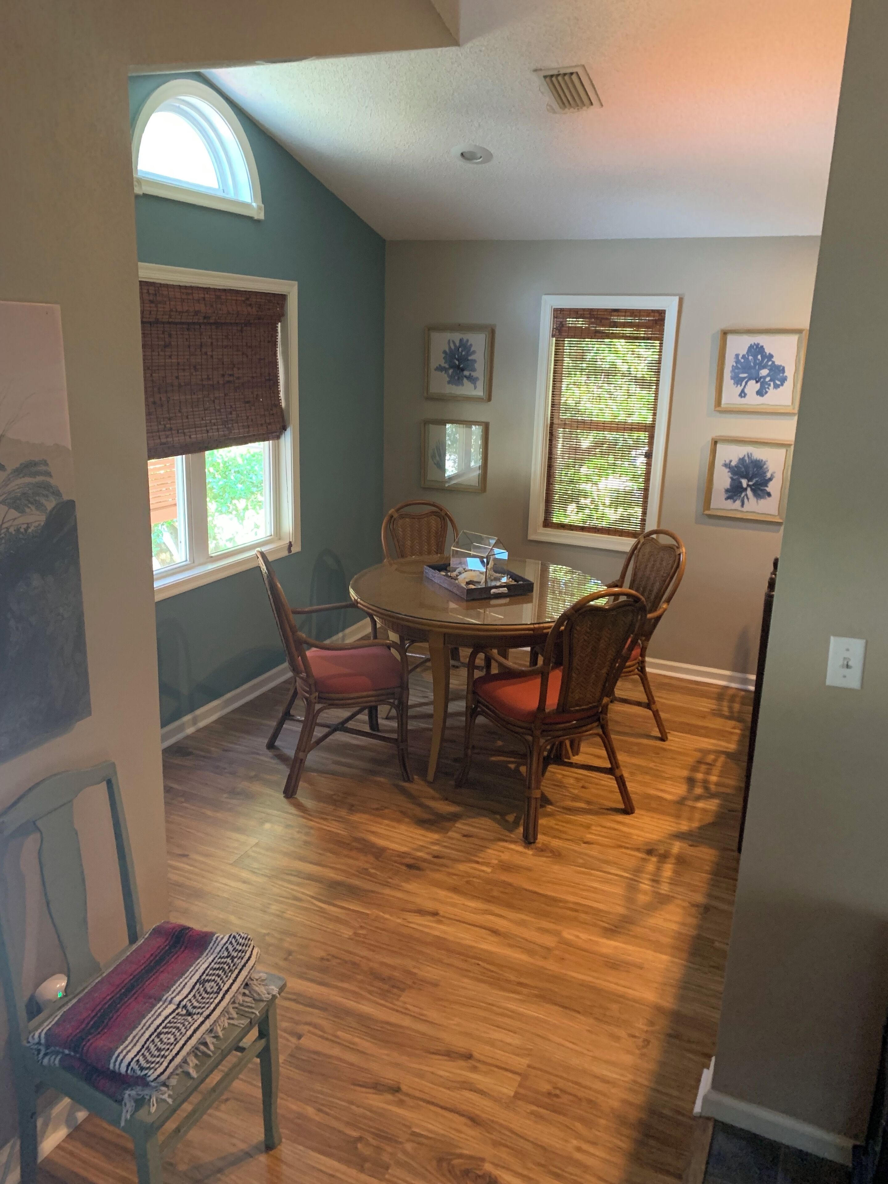 A dining area with a round table, four chairs, and a bench. There are two windows with blinds, a ceiling vent, and framed artwork on the walls.