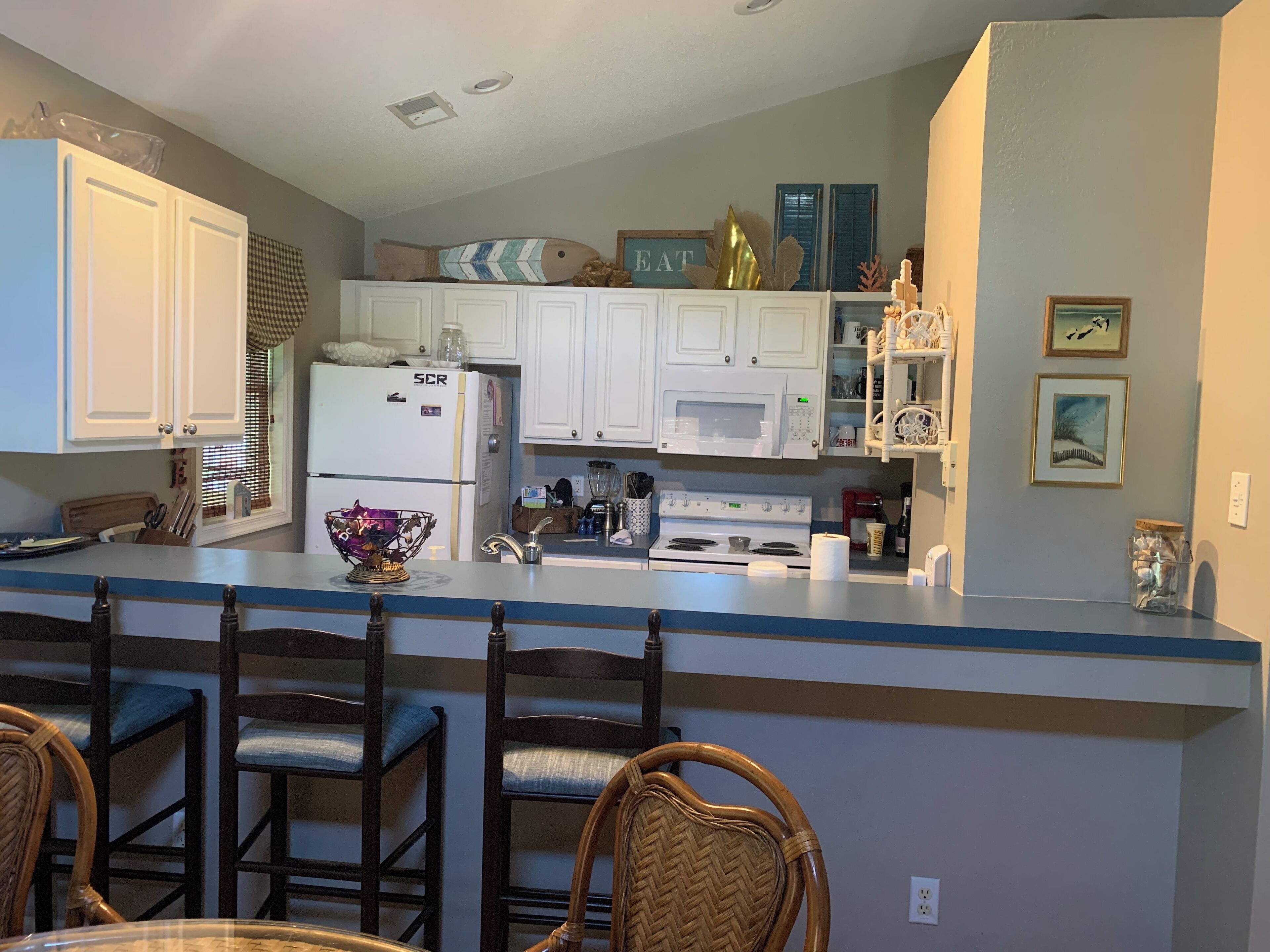 A kitchen with white cabinets, a refrigerator, a microwave, and a stove. There are wicker chairs and a table in the dining area.