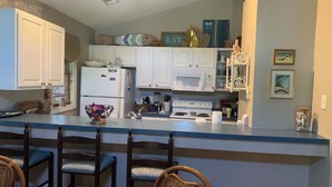 A kitchen with white cabinets, a refrigerator, a microwave, and a stove. There are wicker chairs and a table in the dining area. - Private, Spacious Beach Retreat with Fenced in Shaded Yard (Kill Devil Hills)