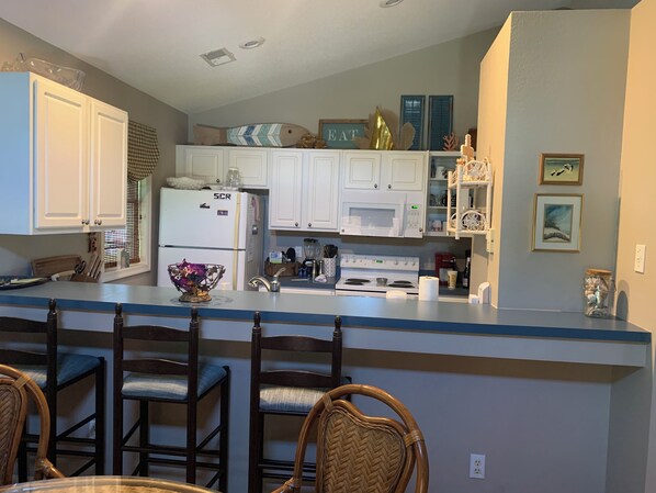 A kitchen with white cabinets, a refrigerator, a microwave, and a stove. There are wicker chairs and a table in the dining area. - Private, Spacious Beach Retreat with Fenced in Shaded Yard (Kill Devil Hills)