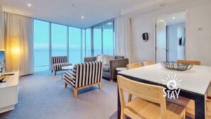 Una habitación de hotel moderna con un ventanal que ofrece vistas al mar, un área de comedor con mesa y sillas, y una sala de estar con sofás y sillones.