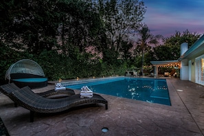 A swimming pool with lounge chairs and a covered seating area, surrounded by trees and a house.