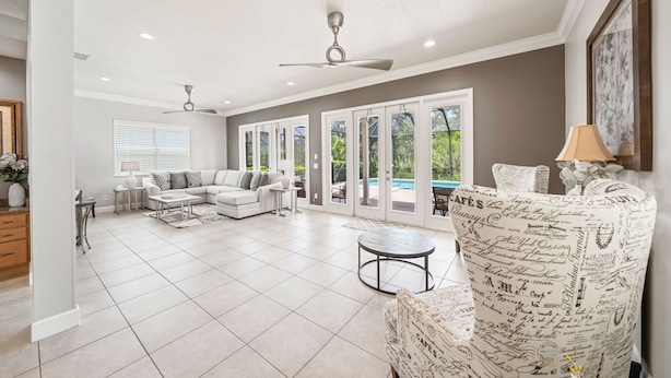 Ein geräumiges Wohnzimmer mit einem weißen Sofa, einem gemusterten Sessel und einem Couchtisch. Zwei Deckenventilatoren, ein Fenster mit Jalousien und Blick auf einen Pool sind vorhanden.