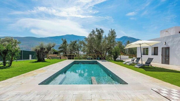 A rectangular swimming pool with a clear blue water surface, surrounded by a paved deck. There are two lounge chairs with umbrellas, a white building with a glass door, and a view of mountains in the background. - Cortijo Villa Maroma Órgiva by Ruralidays (Granada)