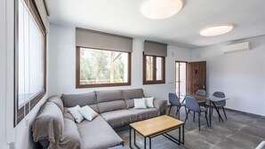 A modern living room with a sofa, a dining table, and chairs. There are large windows providing natural light and a view of trees outside. - Cortijo Villa Maroma Órgiva by Ruralidays (Granada)