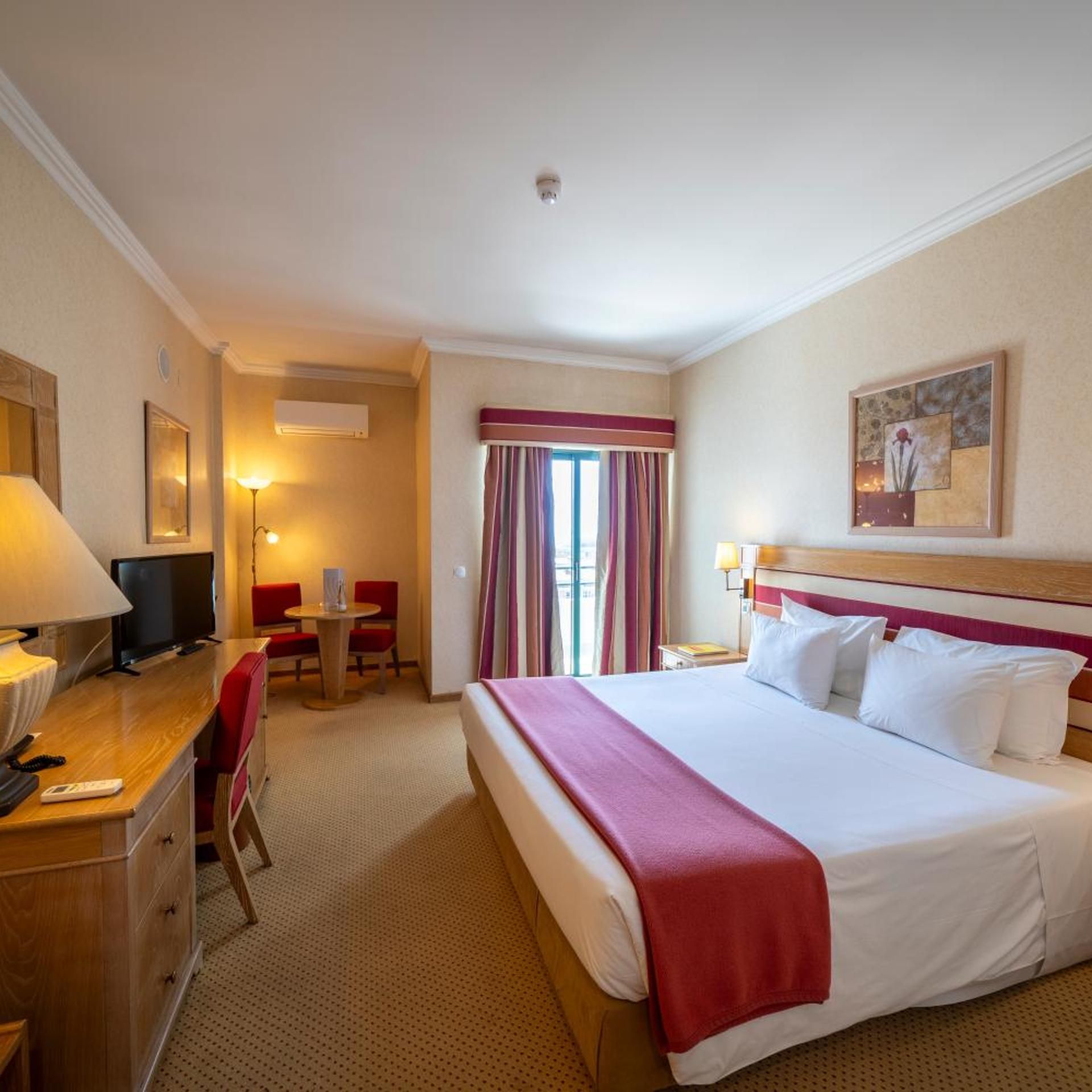 A hotel room with a bed, a desk with a TV, a chair, a small table, and a painting on the wall.