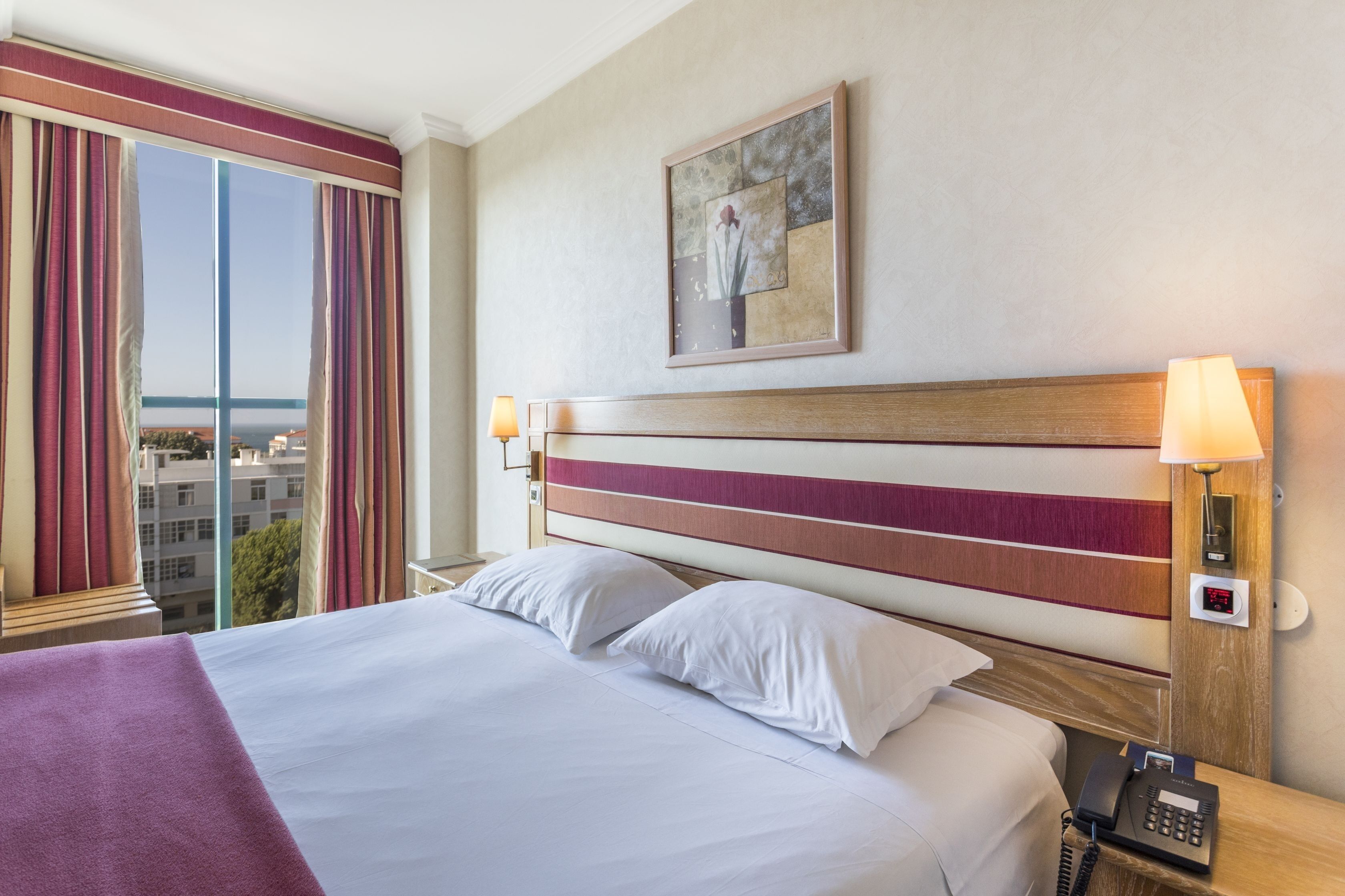 Ein Hotelzimmer mit einem großen Bett, zwei Nachttischlampen, einem Telefon, einem Gemälde an der Wand und einem Fenster mit Vorhängen.