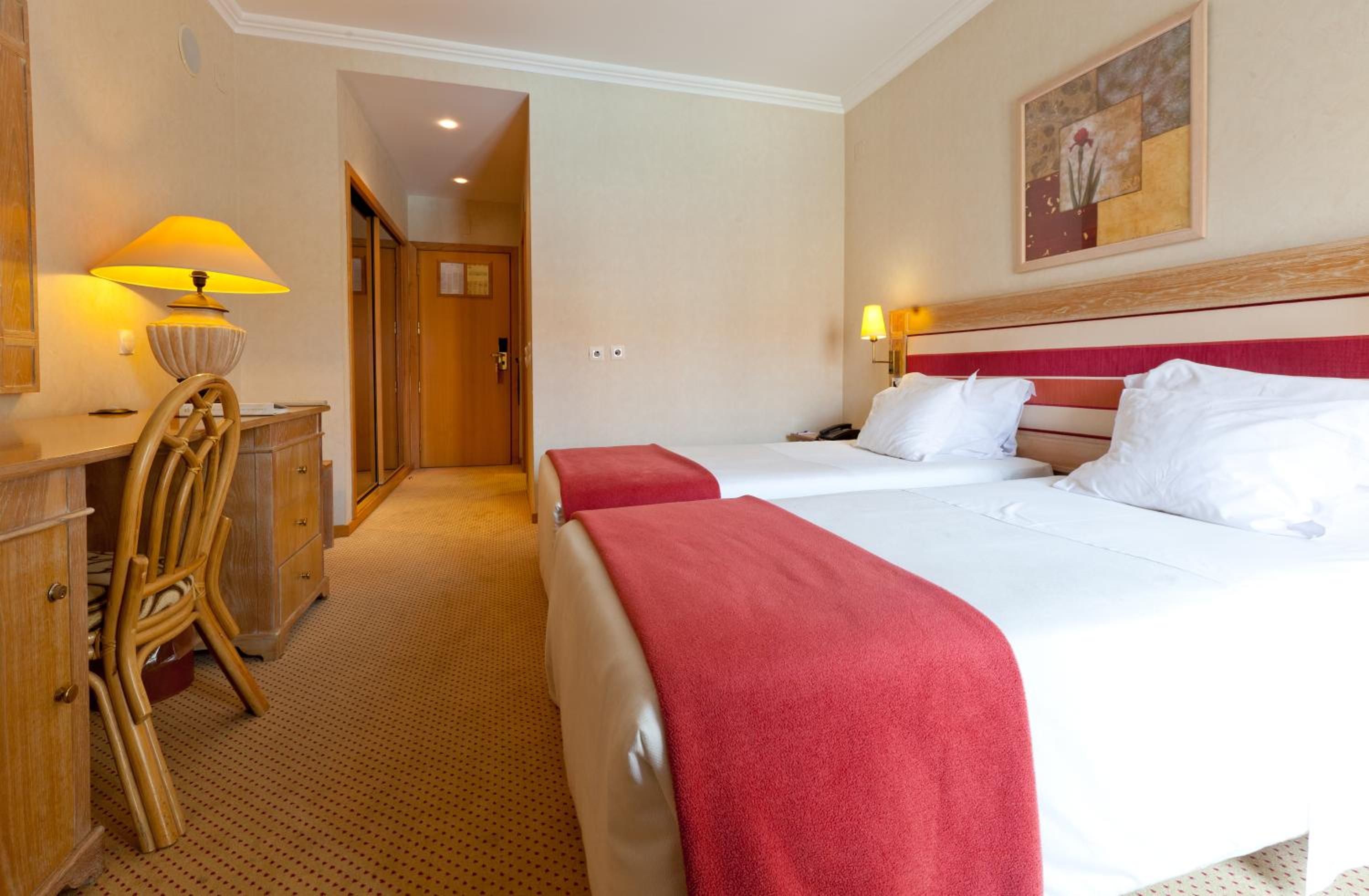 Habitación de hotel con dos camas, un escritorio con lámpara, una silla y un cuadro en la pared.