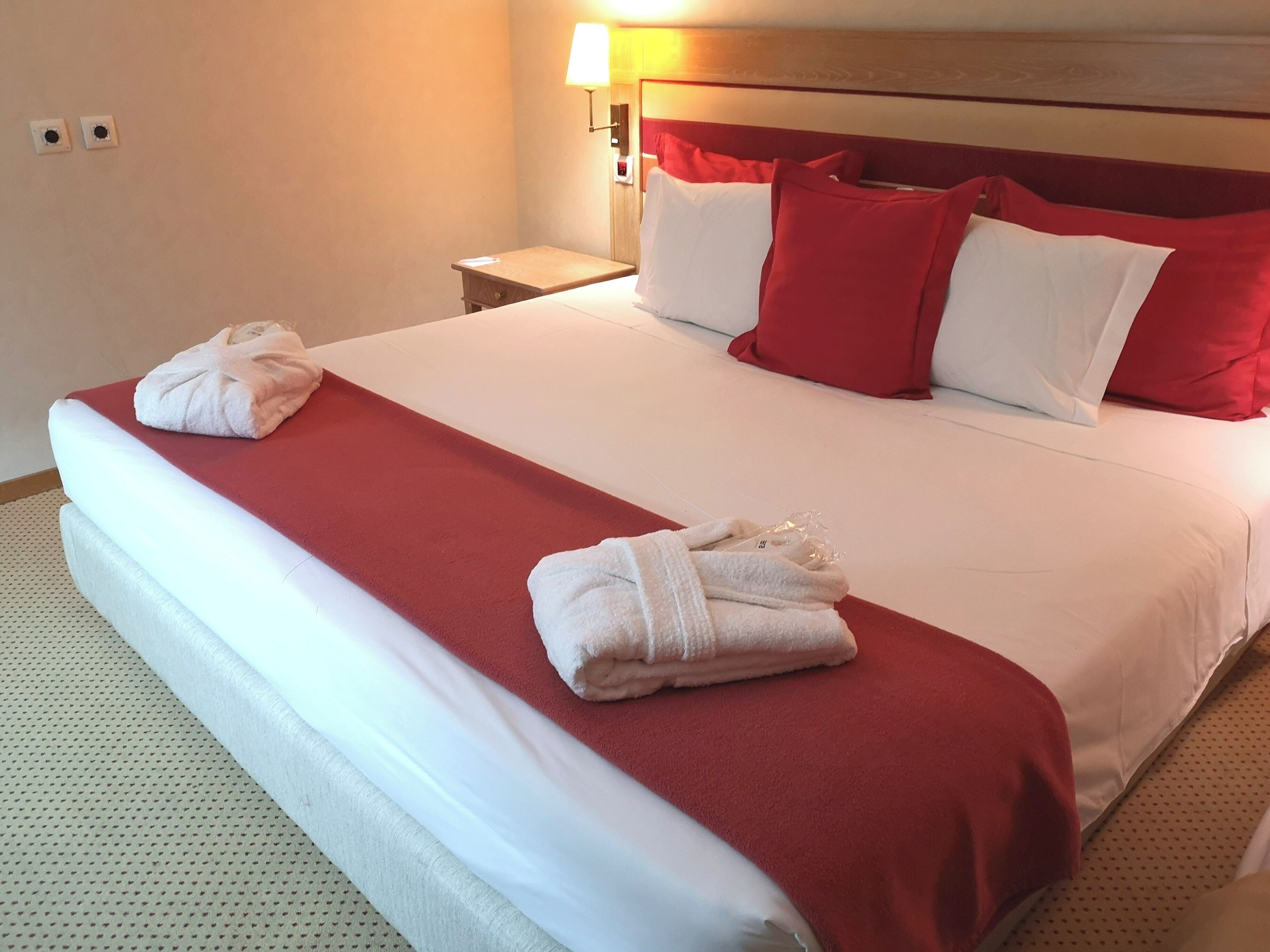Un lit bien fait, avec une literie rouge et blanche, deux serviettes pliées posées sur le lit, une table de chevet avec une lampe, et un mur avec deux interrupteurs.