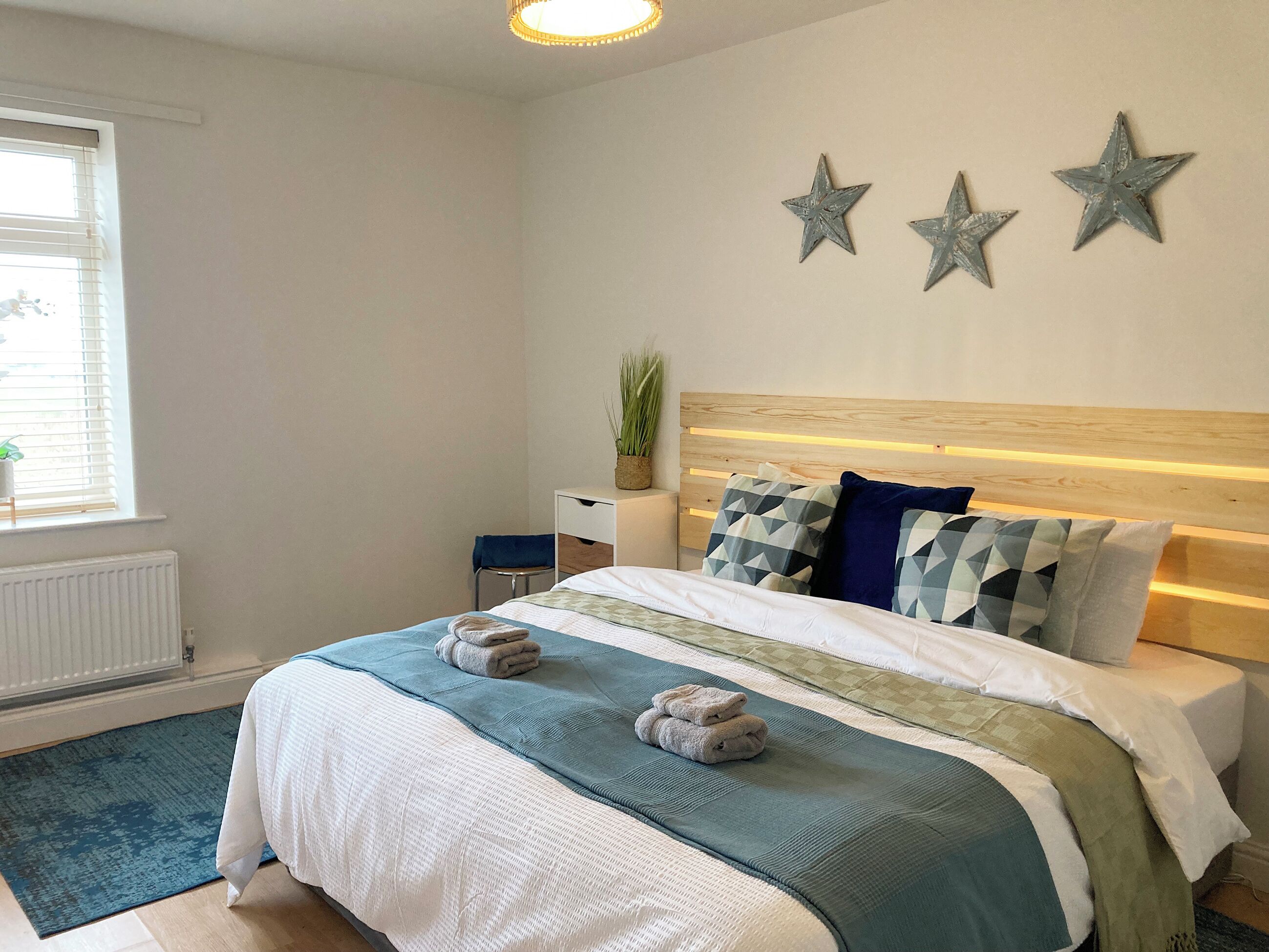 A neatly arranged bedroom with a bed, bedside tables, a radiator, and a window with blinds.