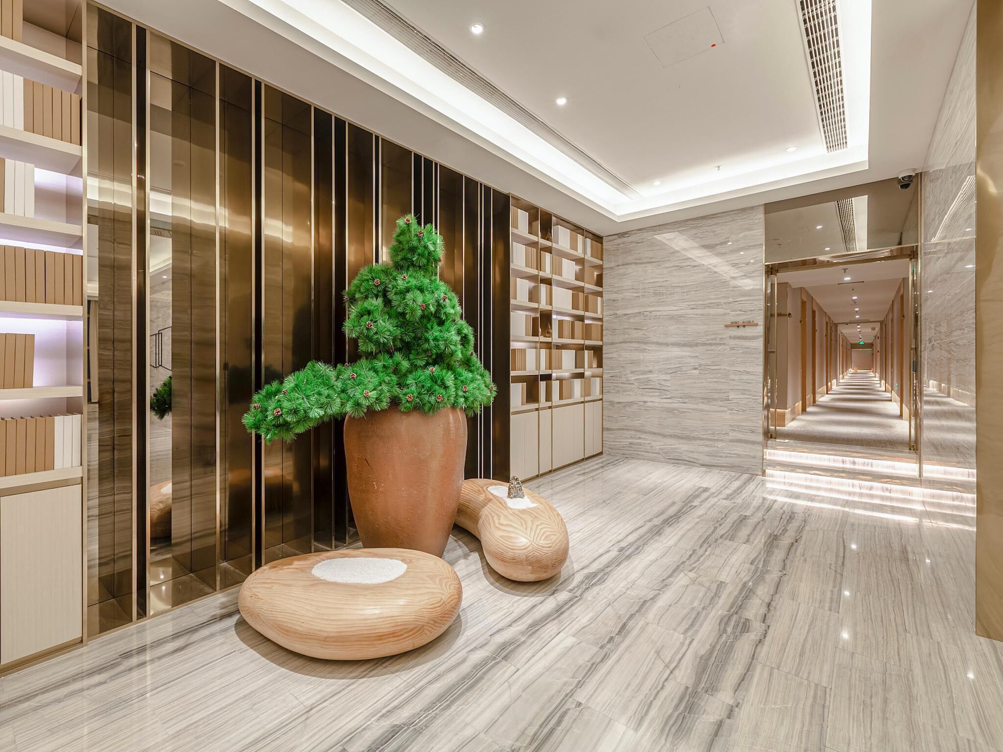 A modern hotel corridor with a large potted plant, wooden furniture, and a marble floor.