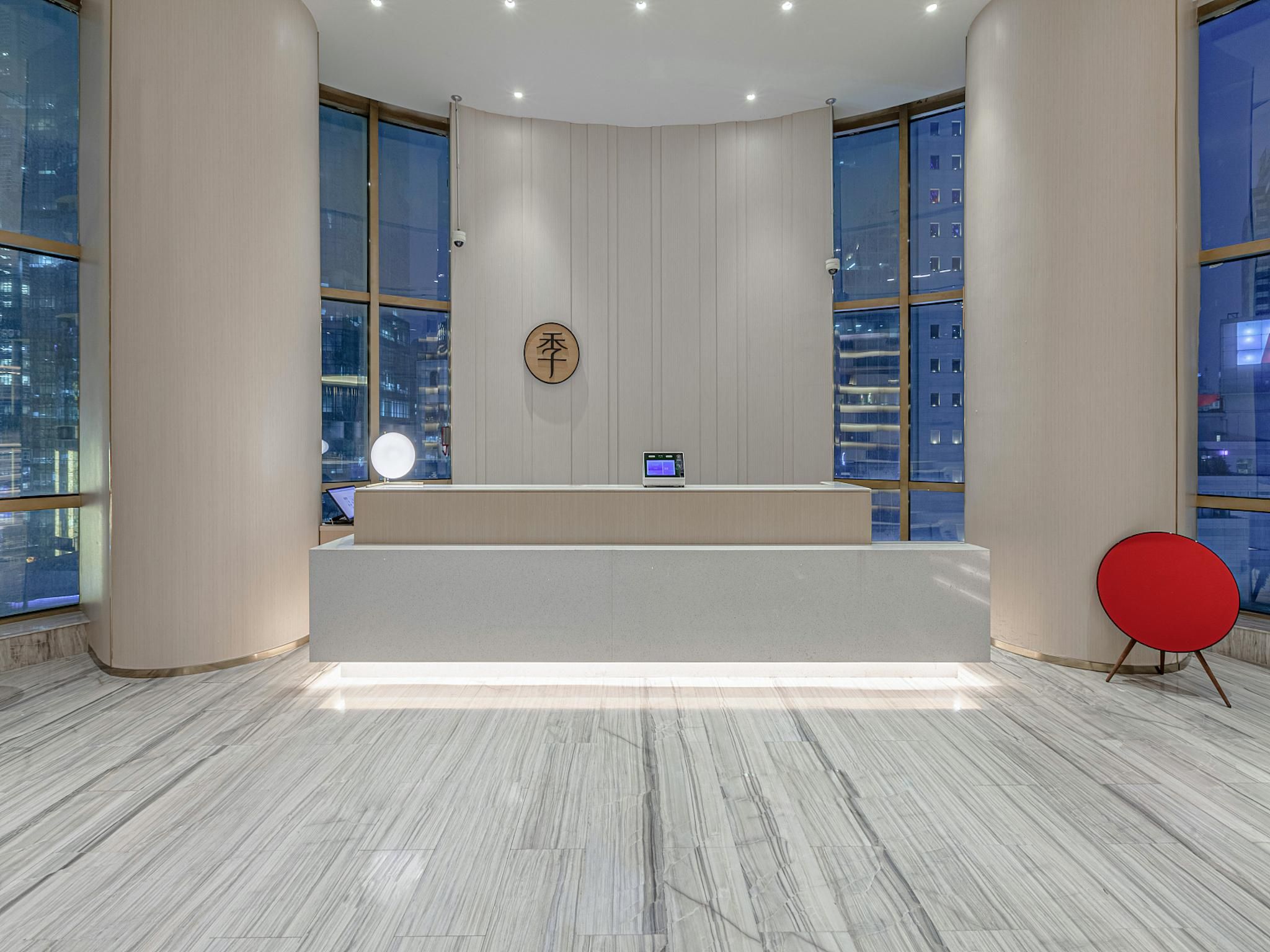 Un comptoir d’accueil avec un ordinateur, un panneau rond et une chaise rouge, dans un hall moderne doté de grandes fenêtres et d’un sol en marbre.