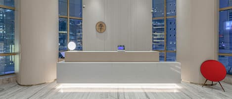 Un comptoir d’accueil avec un ordinateur, un panneau rond et une chaise rouge, dans un hall moderne doté de grandes fenêtres et d’un sol en marbre.