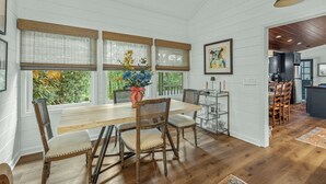 A dining area with a wooden table and wicker chairs, large windows with blinds, and a view of greenery outside. - Linger Longer: One-Level Luxury Home w/ Screened Porch, 2 Fireplaces, Firepit (Highlands)