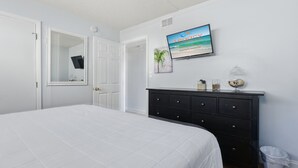 A bedroom with a bed, a dresser, a television mounted on the wall, and a trash can.