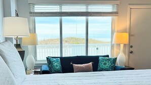 Habitación de hotel con una cama, dos lámparas de noche, una ventana con vista a la playa y una puerta con un cartel de precaución.