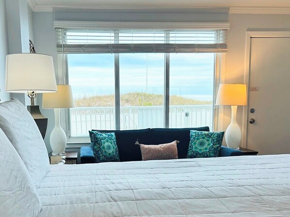 A hotel room with a bed, two bedside lamps, a window with a view of the beach, and a door with a caution sign.