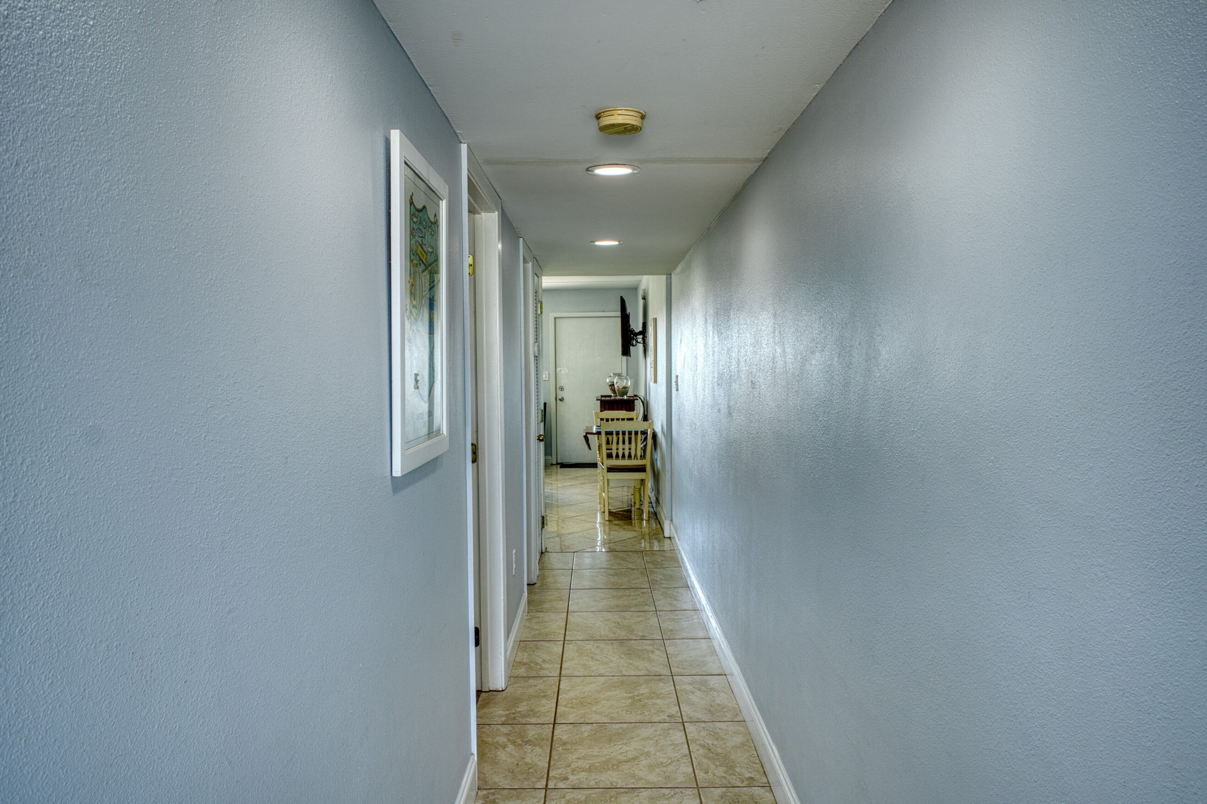 A narrow hallway with light blue walls, a tiled floor, and a wooden chair in the distance.