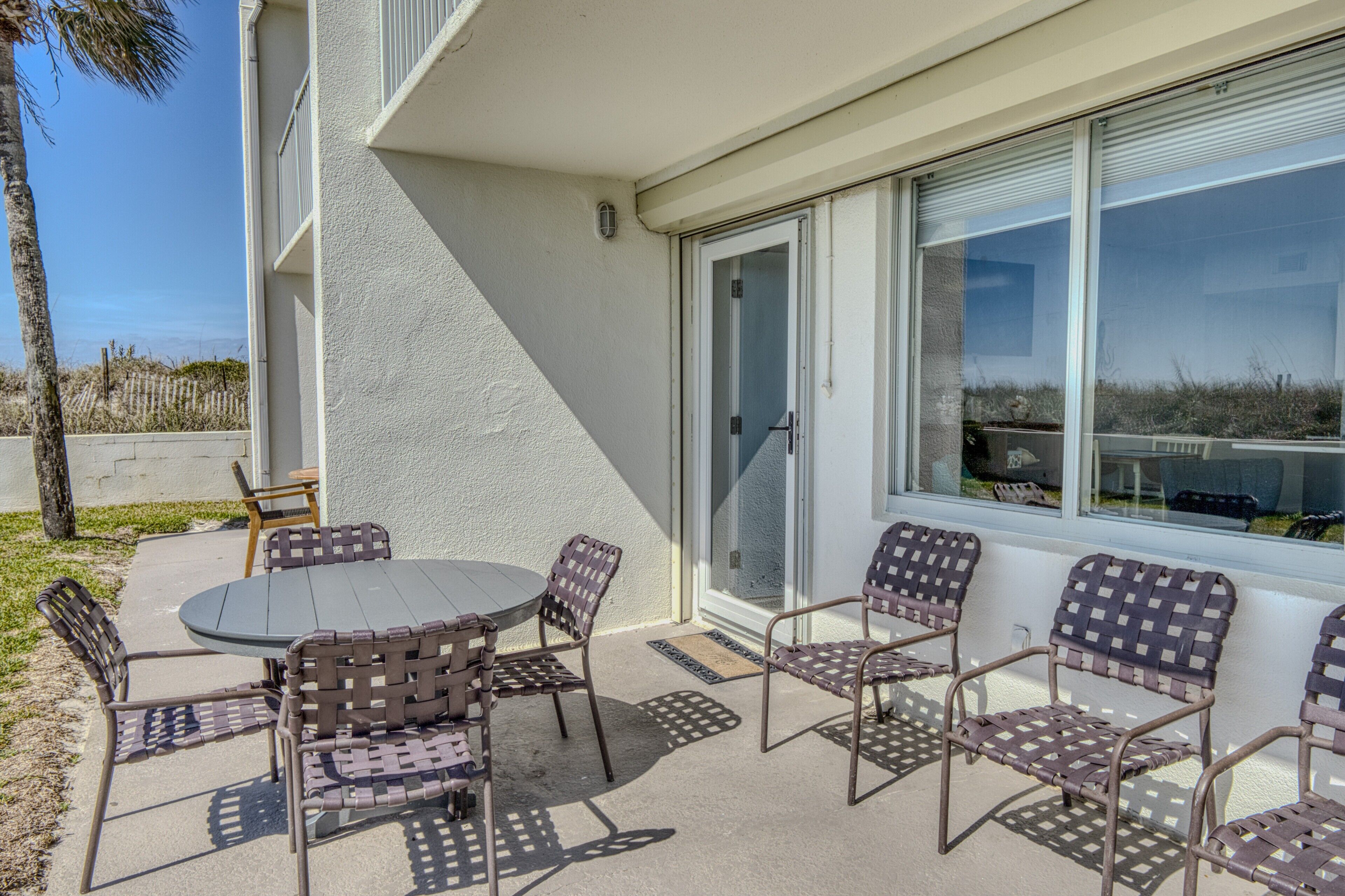 A patio with a round table and six wicker chairs, a sliding glass door, and a view of the beach.