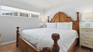 A bedroom with a wooden bed, white bedding, a nightstand with a lamp, and a window with a view of buildings.